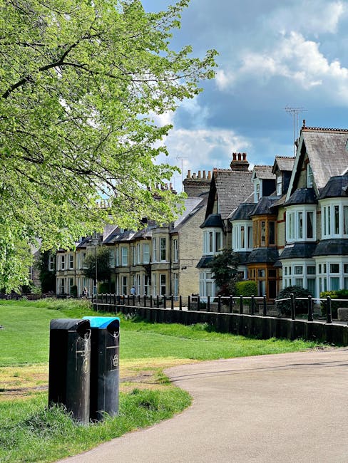 A residential street scene featuring a row of traditional terraced houses with bay windows, pitched slate roofs, and brick facades in various shades of beige, gray, and white. In the foreground, there are two black bin collection points situated on a grassy verge next to a dirt pathway. A large, leafy tree with vibrant green foliage extends across the upper left corner of the image, providing partial shade to the area. The sky is bright with scattered white clouds, suggesting a clear day. The scene appears to be part of a peaceful suburban neighborhood, possibly relating to private waste disposal or rubbish collection services, as indicated by the presence of designated waste receptacles. The greenery and well-maintained appearance of the houses and surroundings contribute to an orderly environment, aligning with the context of rubbish removal and in-situ waste management services, such as those provided by Rubbish Removal Swiss Cottage.