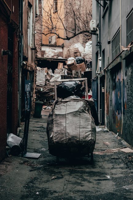 A narrow urban alleyway littered with discarded rubbish and clutter, featuring a large wheeled garbage bag in the foreground, made of worn, light-colored fabric with visible creases and dirt marks. Behind it, there are overflowing piles of mixed waste materials including cardboard boxes, plastic bags, and miscellaneous debris, stacked haphazardly on a makeshift wooden platform at the back of the alley. The alley is flanked by tall, weathered walls on either side; the left side shows dark, rusted metal siding, while the right side has grey painted surfaces with graffiti markings. Overhead, leafless tree branches extend across the scene, and a beige building with windows is visible further in the background. The ground appears damp, with patches of dirt, scattered trash, and a black trash can on the left side. This scene reflects a typical setting where independent rubbish collection or on-site waste clearance might be required, indicating the need for professional rubbish removal services in compliance with local waste disposal regulations, as provided by Rubbish Removal Swiss Cottage.