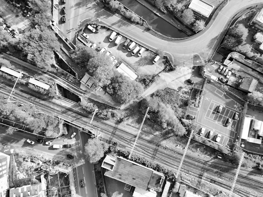 An aerial black-and-white photograph capturing a section of an urban environment featuring railway tracks running horizontally across the centre of the image, with overhanging power lines supported by tall poles. To the left of the tracks, there is a small outdoor parking area with numerous parked cars, adjacent to a building with a flat roof. Behind the parking lot, a narrow path leads to a cluster of trees and shrubs. In the top right corner, a paved road curves and intersects with other streets, with several vehicles visible on the road. To the right of the railway, a basketball court with painted lines and a hoop is seen, with a few cars parked nearby. The surrounding area includes residential and commercial buildings, some with pitched roofs and others with flat roofs, embedded within a landscape of trees, bushes, and open spaces. The overall scene illustrates an urban space with infrastructure and vegetation, aligning with typical locations where private rubbish removal and on-site clearance services by firms such as Rubbish Removal Swiss Cottage might be employed to manage waste from residential or commercial properties. The environment appears well-lit with natural sunlight, emphasizing the textures of surfaces and greenery.