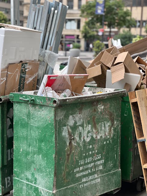 A large, green, metal waste skip positioned on a paved outdoor area, filled with discarded packaging materials, cardboard boxes, and other miscellaneous rubbish. The bins exhibit signs of rust and dirt, with some surface scratches and paint peeling slightly. In the background, there are urban surroundings including trees, multi-storey buildings, and storefronts, indicating an outdoor street setting. The sky is overcast, providing diffuse natural lighting that highlights the textures of the rubbish and the skip surfaces. The scene suggests an off-site waste collection or cleanup activity, consistent with private or independent rubbish removal services, such as those offered by Rubbish Removal Swiss Cottage, providing an alternative to local council bin collection for larger or bulk waste disposal needs.