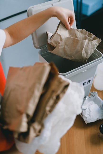 A person depositing a crumpled brown paper bag into a grey rubbish bin with a partially visible lettered label, situated indoors on a wooden surface. The person's hand is grasping the bag, which is lined with a black plastic bag inside the bin. In the foreground, another crumpled brown paper bag is held in the person's other hand, with some paper waste nearby on the surface. The background includes a white wall and a glimpse of a black object, suggesting a domestic or small commercial setting. The scene highlights the process of manual waste disposal, reflecting practices associated with independent rubbish collection or on-site waste handling. The lighting is natural, and the textures of the paper, plastic, and wood are clearly visible, emphasizing the ordinary act of rubbish disposal within a household or small business context, as managed by local waste services such as Rubbish Removal Swiss Cottage.